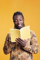 Intellectual african american man relaxing reading novel book, enjoying learning new information standing in studio over yellow background. Cheerful person studying literature, liking genre and plot photo