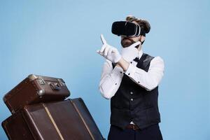 Doorman employee uses vr glasses on camera and posing next to pile of suitcases, having fun with modern 3d interactive vision headset. Bellboy exuding professionalism and modernization. photo