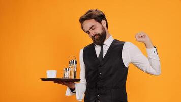 Overworked waiter feeling tired on camera, yawning and stretching on duty. Experienced employee in suit and tie feeling tired and almost falling asleep in studio, poses in yellow studio. photo