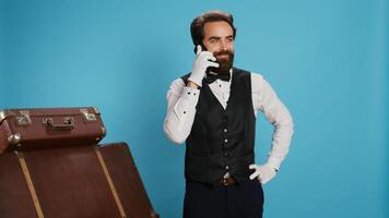 Doorman talks on phone call with hotel guests, making room reservations against blue background. Professional bellhop chatting with visitors on camera and accepting remote bookings. photo