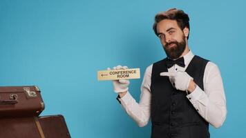 Elegant bellboy has conference room symbol in hand, showcasing wall pointer to lead hotel guests to all area on the premises. Experienced porter posing with an indicator wall sign. photo