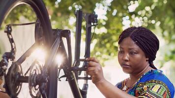 determinado negro mujer pruebas y inspeccionando bicicleta rueda y pedales en yarda para verano ciclismo. africano americano hembra ciclista con caucásico hombre al aire libre reparando bicicleta partes utilizando trabajo herramientas. foto