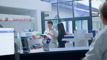 Asian woman in need of prescription medication informations in apothecary, asking cashier for help who redirects her to chemist checking pharmaceutical products inventory photo