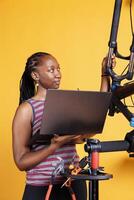 Youthful black lady using guidelines from electronic device to carefully examine and fix her broken bike. Sporty woman uses laptop and specialized tools to examine modify and repair bicycle. photo