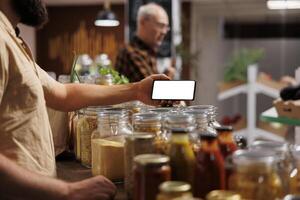 Man in zero waste shop using mockup mobile phone to analyze products. Vegan customer thoroughly checking local supermarket food items are suitable for his diet with isolated screen smartphone photo