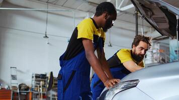 Mechanics in auto repair shop working together on fixing car, using tablet and laptop. Coworkers collaborating on servicing broken vehicle, checking for defective components using electronic devices photo