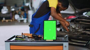 Mechanic in car service uses wrench to replace parts inside vehicle with green screen tablet in front. Expert utilizes professional tools to adjust malfunctioning automobile next to mockup device photo