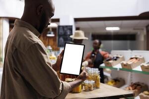 African American guy browses the bio-food items, jars, and foods on shelves while carrying a phone tablet with a chromakey template. Black man grasping a digital device with a white screen. photo