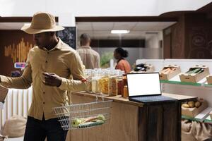 While African American clients browse the store a wireless computer with a blank mockup template is placed on wooden counter. side view shot of a laptop showing an isolated white screen for adverts. photo