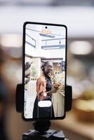 African American store owner uses cell phone and selfie stick for vlogging, promoting sustainable, healthy and natural items. Organic, fresh food in glass jars, appealing to eco-conscious customers. photo