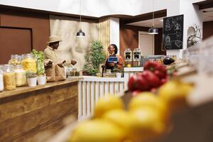 African American man with hat on appears to be completing a purchase at the cashier desk in a grocery store. The shop is eco friendly and promotes green and sustainable lifestyle. Focus on background. photo