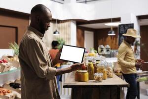 While carrying tablet with chromakey template, African American guy checks eco friendly items, jars and foods on shelves. Male customer horizontally grasping a device displaying isolated white screen. photo