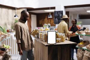 In an eco friendly store, a detailed look of a tablet with an isolated chromakey mockup template is shown. Close-up of digital device with blank white screen display, positioned near food glass jars. photo