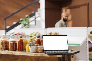 Wireless computer displaying white screen in bio shop filled with freshly harvested fruits and vegetables. Digital laptop showing isolated chromakey mockup template in zero waste store. photo