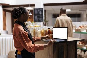 In a grocery store, African American female vendor examines the products with laptop displaying blank chromakey template. The eco friendly shop shows a variety of reusable containers and glass jars. photo