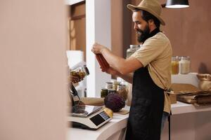 Caucasian male shopkeeper, who wears a stylish hat and black apron, stands behind the checkout counter holding a jar filled with pasta sauce. Vendor analyzing glass container at cashier desk. photo