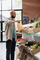 Middle eastern man explores modern eco friendly grocery store, browsing a variety of fresh, organic products with sustainable packaging. Male customer putting fresh potatoes in brown paper bag. photo