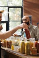 Middle Eastern customer looks at glass bottle filled with organic sauce being held by storekeeper. Local vendor providing male client with bulk natural sustainable products during an event. photo