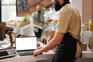 In zero waste convenience store, storekeeper waits to serve customers while using digital laptop with white screen. Caucasian seller using minicomputer displaying isolated chromakey template. photo