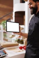 While holding tablet with isolated white screen, male shopkeeper waits to assists customers. Caucasian male seller gripping a digital device horizontally with blank copyspace mockup template. photo