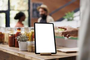 Electronic gadget with white screen is placed on a table filled with various glass jars and bottles in eco friendly grocery store. Focus on tablet that displays isolated chromakey mockup template. photo