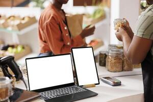 Digital screens on counter showing blank chromakey mockup template intended for eco friendly store advertisement. Tablet and laptop at cashier desk with isolated white screen. photo