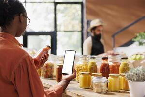 African american customer analyzes glass bottle filled with red sauce while also holding mobile device with white screen in grocery store. Black woman grasping cellphone with blank mockup template. photo