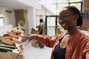 Black female vlogger showcasing various locally grown produce of eco friendly store. African american woman on video call pointing towards freshly harvested fruits and vegetables in grocery shop. photo