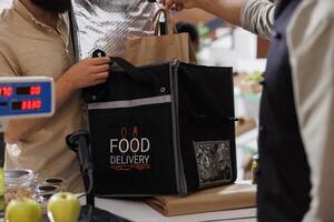 Close-up of the storekeeper placing takeaway into thermal bag for delivery man to send to customers with healthy living. Male courier collecting grocery order at counter of eco friendly store. photo