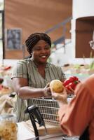 Happy african american female individual shopping for locally grown organic vegetables for healthy recipes. Young consumer giving local vendor the fresh produce for weighing and packaging. photo