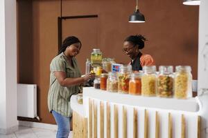 African American customer discusses eco friendly shopping with a store clerk at the checkout while handing over jars of cereal and honey for scanning. Black woman purchasing fresh bio food products. photo