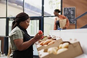 Female storekeeper checking and arranging freshly harvested produce on shelves. Black woman wearing an apron holding and examining red apples in boxes at local convenience store. photo