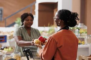 Black woman at the checkout of a zero-waste store, purchasing locally grown eco-friendly vegetables from the storekeeper. Female customer purchasing chemical-free organic groceries. photo