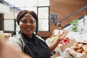 negro mujer vlogging acerca de sano vivo a local tienda de comestibles comercio, demostración bio comida productos tendero utilizando teléfono inteligente a compartir su mensaje como contenido creador y blogger en eco simpático almacenar. foto