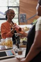 An African American woman sells freshly harvested organic produce to a customer in a grocery market. Female vendor receiving bottled sauce from smiling customer a checkout counter. photo