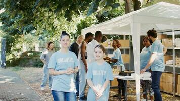 Portrait of a woman and daughter taking part in food bank program to fight hunger. Blue-shirted Caucasian volunteers are ready to help less fortunate while glancing into camera. Handheld shot. photo