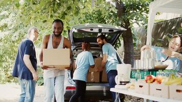 An outdoor non-profit food drive program to aid the poor needy and homeless people. Young multiethnic volunteers giving donation boxes from the car to the less privileged. Handheld shot. photo