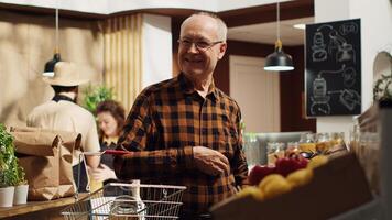 Portrait of smiling old client in zero waste supermarket using shopping basket to purchase bulk items in reusable glass containers. Elderly man in local grocery shop buying pantry staples photo