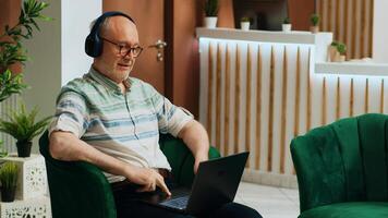 Elderly man watching movie on laptop and enjoying online streaming services at hotel reception lobby. Guest using device to keep up with favorite tv show, waiting for check in process. photo