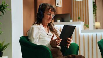 Retired woman using tablet in lobby, watches movie on streaming services while she waits to see room reservation. Old person enjoying modern technology with device and headset. Handheld shot. photo