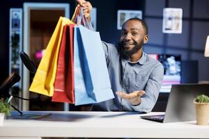 africano americano hombre rodaje Moda vlog, participación compras pantalones en mano, demostración suscriptores compras él recientemente consiguió. Internet espectáculo anfitrión presentación espectadores nuevo adquisiciones, rodaje él mismo a hogar foto