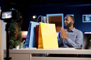 Influencer holding colorful shopping bags in hand, showing subscribers purchases he recently got. Content creator presenting fanbase new acquisitions, filming himself in apartment photo