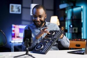 Smiling BIPOC artist filming DJ mixing tutorial in recording studio, playing with turntables, samples and sound effects. Music production content creator showcasing audio equipment to audience photo
