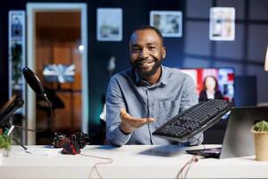 Influencer in home studio using recording tech to capture live broadcast, reviewing mechanical keyboard. BIPOC online show host filming electronics haul, presenting computer peripheral to viewers photo