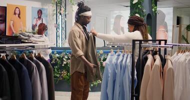 Couple in thrift shop looks at clothes, trying to find well fitting special occasion suits. Woman satisfied by cheap and high quality blazers in SH clothing store, giving ok hand sign approval gesture photo