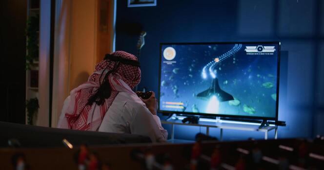 Muslim gamer plays intense classic arcade space shooter videogame, shooting asteroid using laser beams. Arabic man relaxing at home using high tech gaming system to solve missions in singleplayer game photo