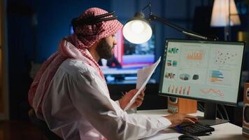 Arab teleworker checking analytics statistical data set while remotely working in stylish apartment. Business manager optimizing key performance indicators with tv as background noise photo