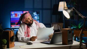 Muslim student at home in telephone call with school colleague discussing about finance class lesson and doing homework tasks together. Arab man at home desk looking over webinar notes photo