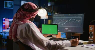 Muslim worker in apartment office holds chroma key tablet while looking at business annual report graphs on computer. Professional freelancer with mockup digital device in hand performing job tasks photo