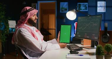 Muslim worker in apartment office holds chroma key tablet, crosschecking data on it with business report graphs on computer. Smiling entrepreneur with mockup digital device in hand performs job tasks photo